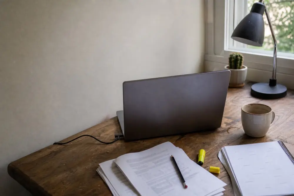 Home-office desk with a laptop, a printed document marked with pencil and a notebook beside a window