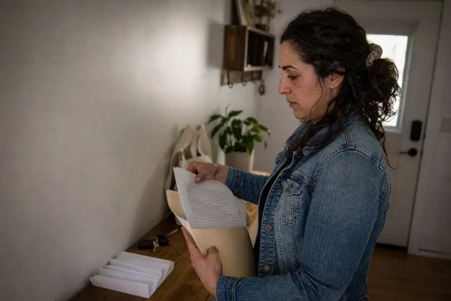 Woman in a home entryway putting printed pages into a manila folder before leaving, keys and a tote bag on the shelf beside her